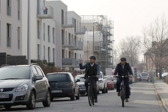 Zwei Fahrradfahrer fahren an Wohnstraße vorbei an parkenden Autos